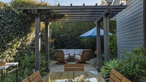 Een houten pergola met een tafel en stoelen, omgeven door groen.