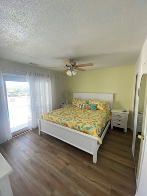 Une chambre à coucher comprenant un lit, un ventilateur de plafond, une table de chevet, une commode et une fenêtre avec des rideaux.