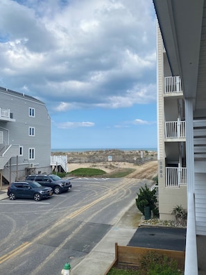 Un parking avec deux voitures, un immeuble doté d’un balcon et, au loin, une plage.