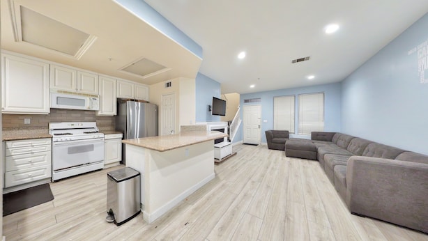 A modern kitchen with white cabinets, a stainless steel refrigerator, and a microwave. There is a gray sectional sofa and a small trash can in the living area. -     Fully Furnished Anaheim Townhome - Utilities Included - Gated Community (Anaheim)