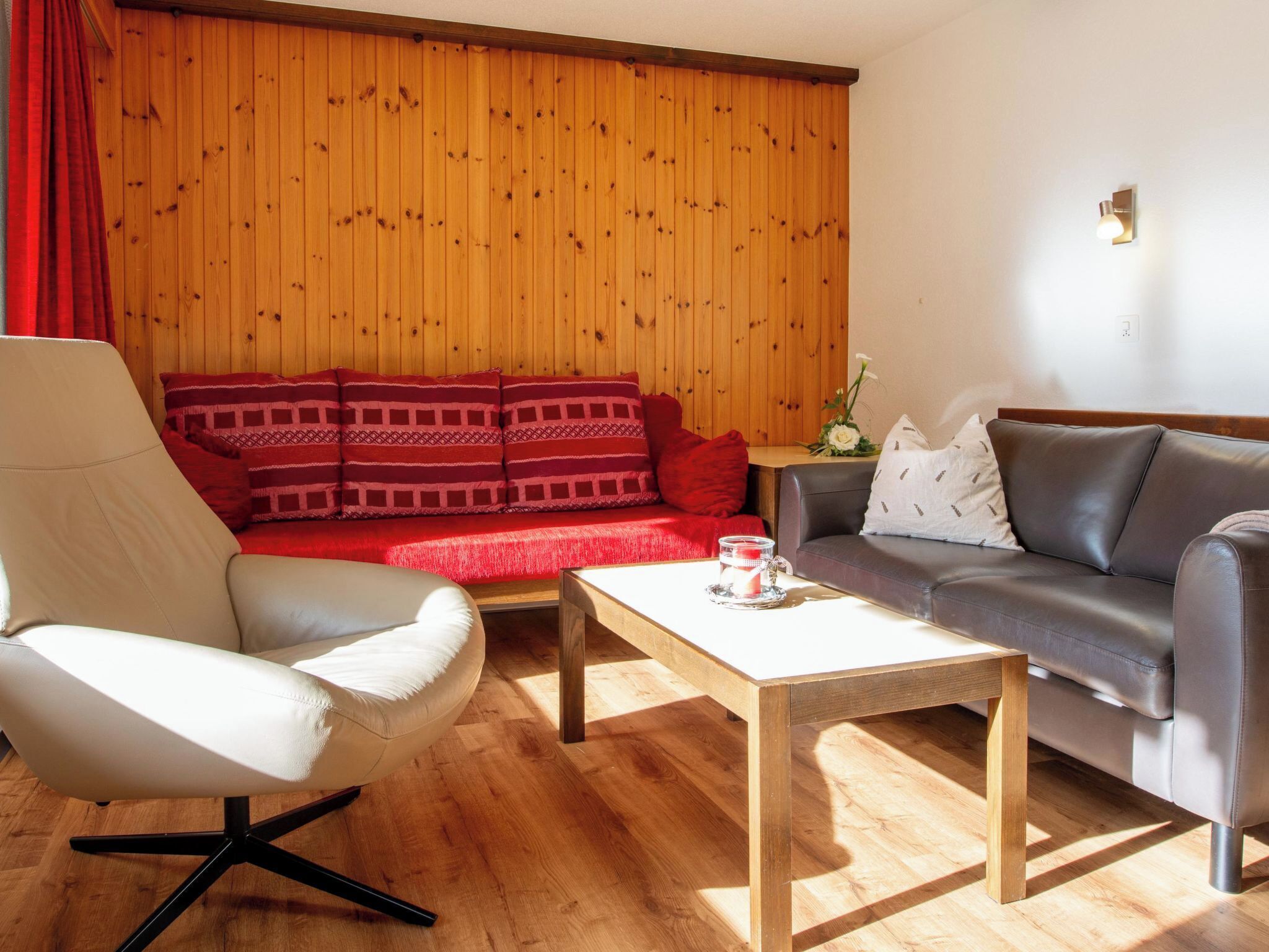 A living room with a wooden paneled wall, a red sofa, a grey sofa, a white armchair, and a wooden coffee table.