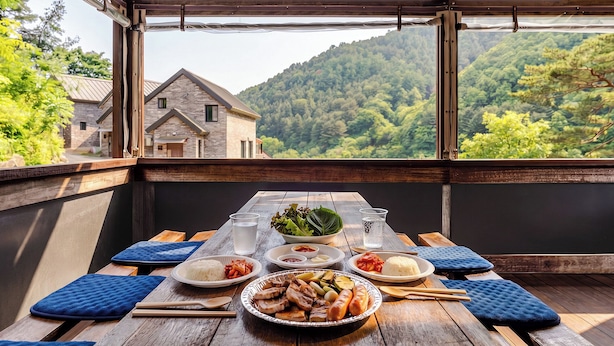 Ein Holztisch mit Speisen, einer Salat-Schüssel und einem Glas Wasser, vor einer Bergkulisse.