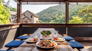 Una mesa de madera con platos de comida, un tazón de ensalada y un vaso de agua, con vistas a una montaña de fondo.