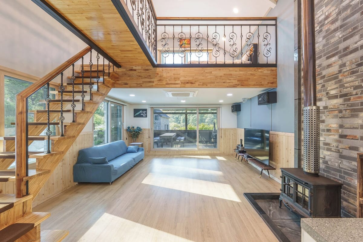 Amplia sala de estar con escalera de madera, un sofá azul, televisor y zona de cocina con pared de ladrillo.