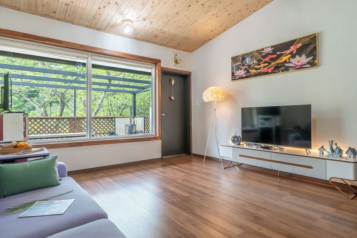Una sala de estar con sofá, televisor, lámpara de pie y un amplio ventanal con vistas a árboles.