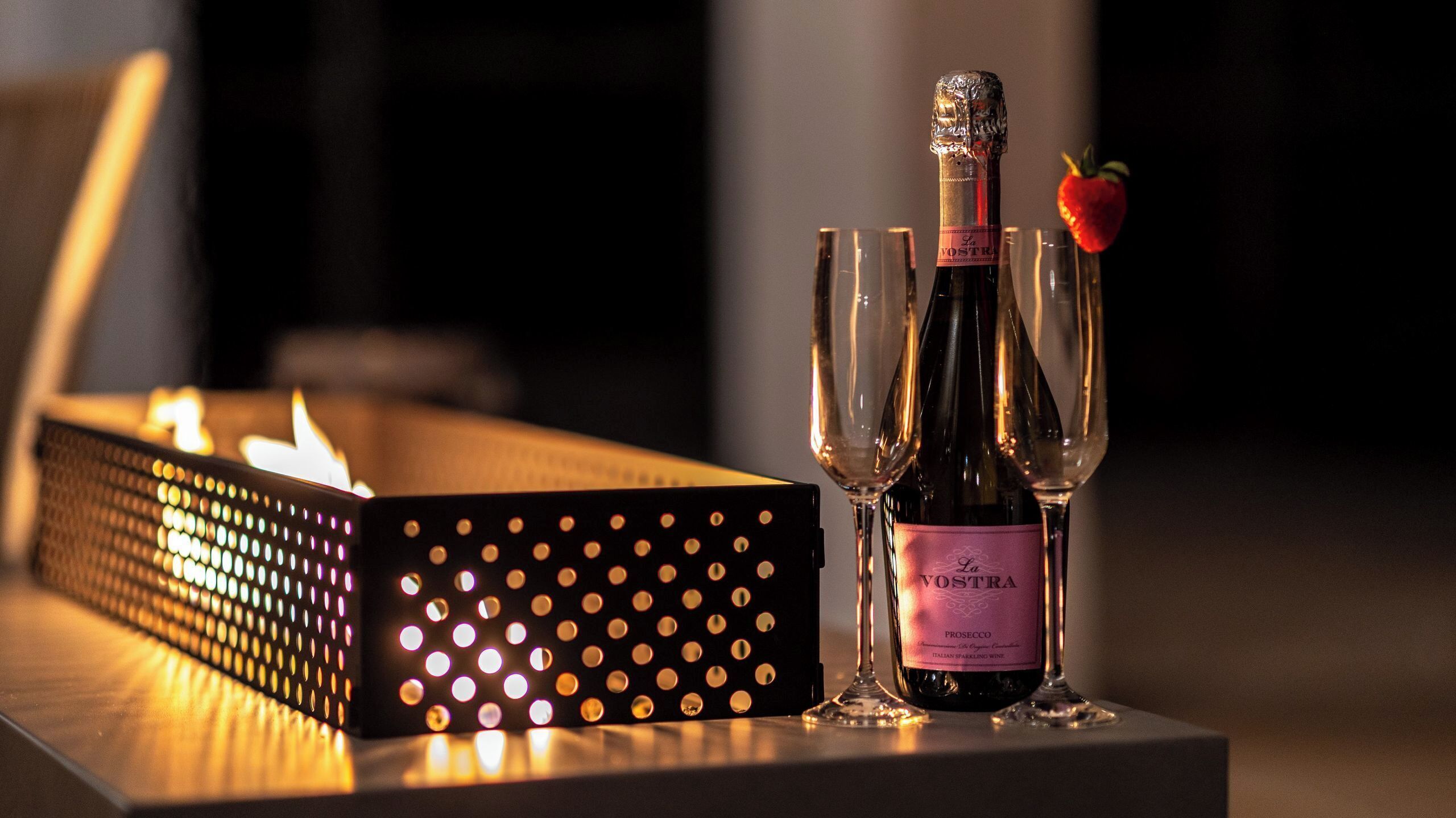 A bottle of La Vostre Prosecco wine with a strawberry garnish, two glasses, and a lit candle on a table.