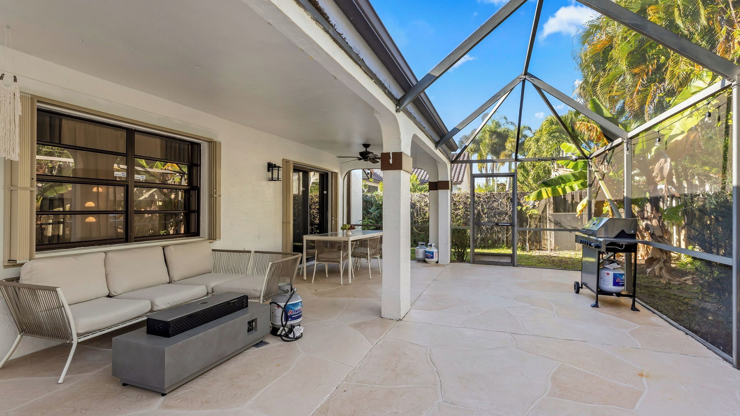 A covered patio with a sofa, table, and grill area, featuring large windows and a view of greenery.