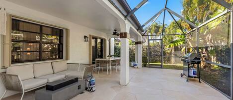 A covered patio with a sofa, table, and grill area, featuring large windows and a view of greenery.