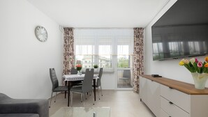 Un salon moderne comprenant un canapé, une table basse en verre, un coin repas avec des chaises et une table, un téléviseur fixé au mur et une fenêtre avec des rideaux.