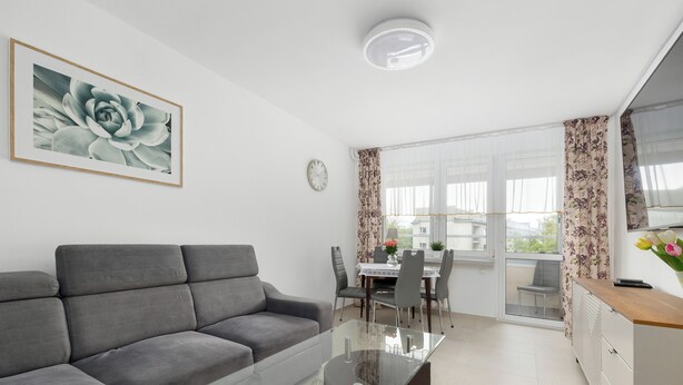 Un salon moderne avec un canapé gris, une table basse en verre, un coin repas comprenant une table et des chaises, et une vue dégagée sur l’extérieur grâce à une grande fenêtre.