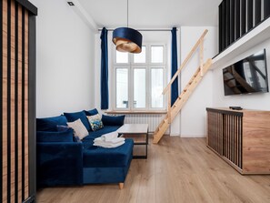 Ein modernes Wohnzimmer mit einem blauen Sofa, einer Holztreppe, einem an der Wand montierten Fernseher und einem Fenster mit Vorhängen.