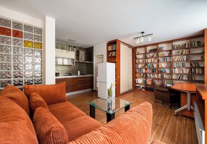 A modern living room with a sofa, a glass coffee table, a bookshelf, and a kitchen area with cabinets and a refrigerator. - Bednarska Royal Route Apartment (Warszawa)