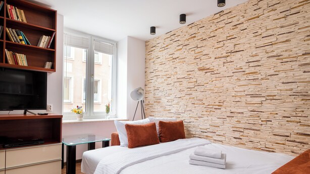 Moderni hotellihuone, jossa on puuseinä, valkoisilla tekstiileillä varustettu sänky, kirjahylly, televisio ja ikkuna, josta on näkymä.