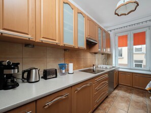 A kitchen with wooden cabinets, a stainless steel range hood, a built-in oven, a coffee maker, a kettle, a toaster, and a window with blinds. - Łódź Spacious Apartment (Łódź)
