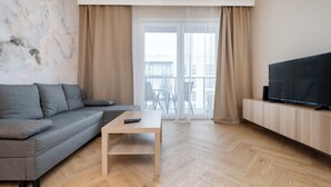 A modern living room with a grey sofa, a wooden coffee table, a TV on a wooden stand, and a large window with curtains.