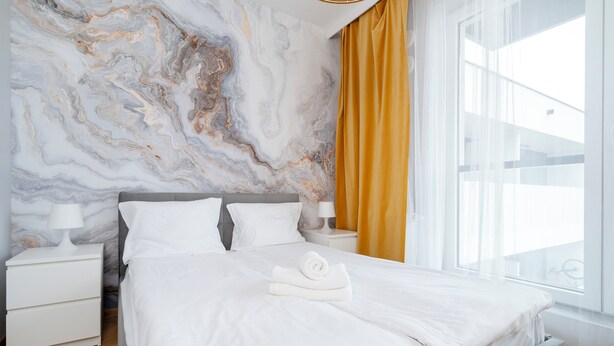 A modern bedroom with a marble wall, a large bed, a nightstand, a lamp, and a window with curtains.