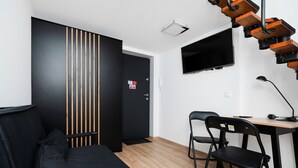 Quarto com sofá preto, duas cadeiras pretas, escrivaninha de madeira, luminária preta e televisão fixada na parede.