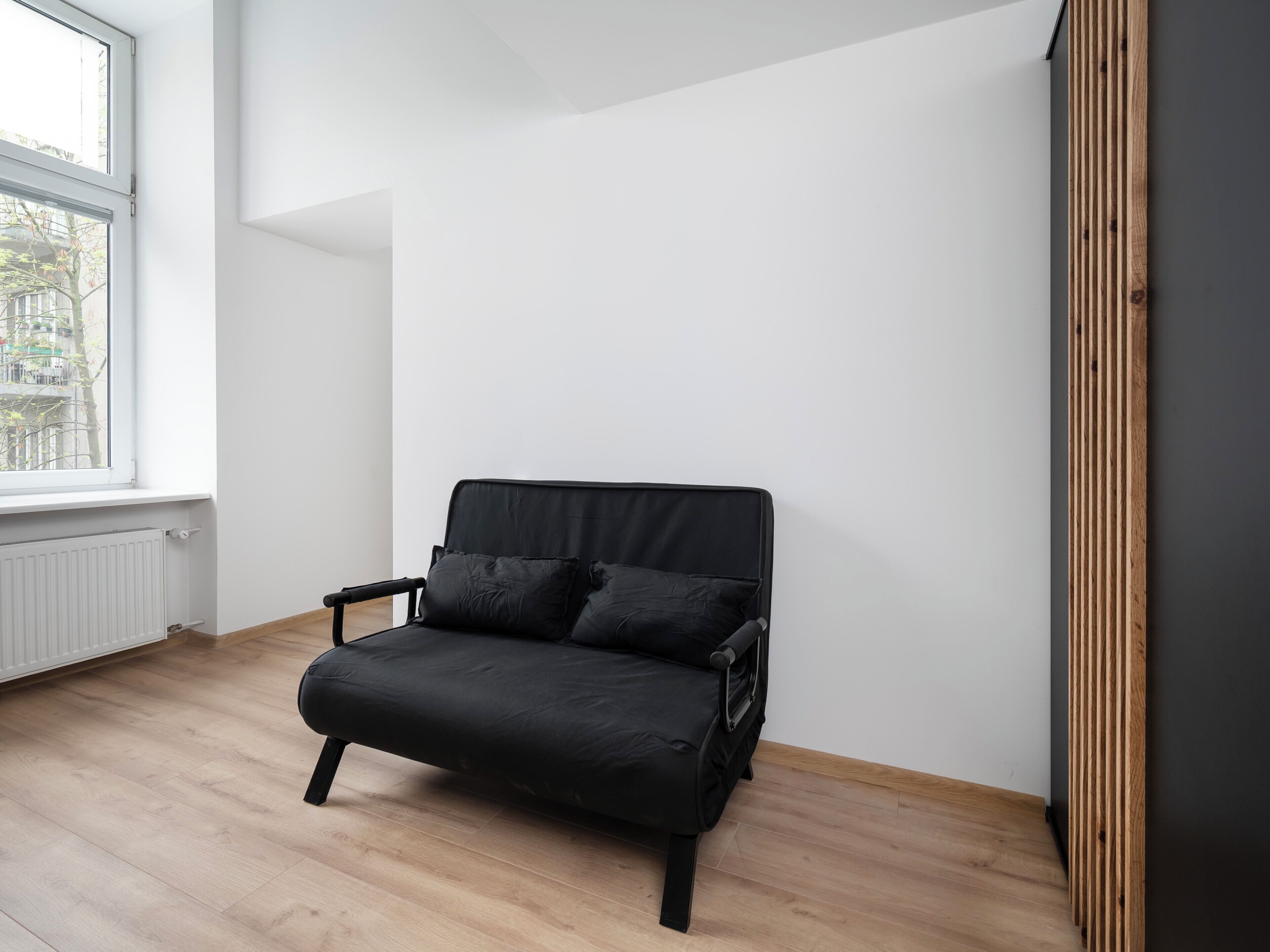 A black chair in a room with a window, white walls, and wooden flooring.