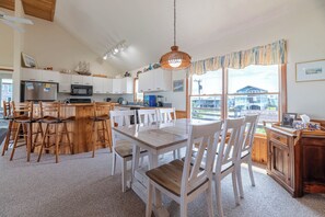 A kitchen with a central island, wooden chairs, a dining table, and a view of the outside. - Ocean Wynns - Nags Head MP 13, 20 Yards to the Beach, Ocean Views, Close to Attractions (Nags Head)