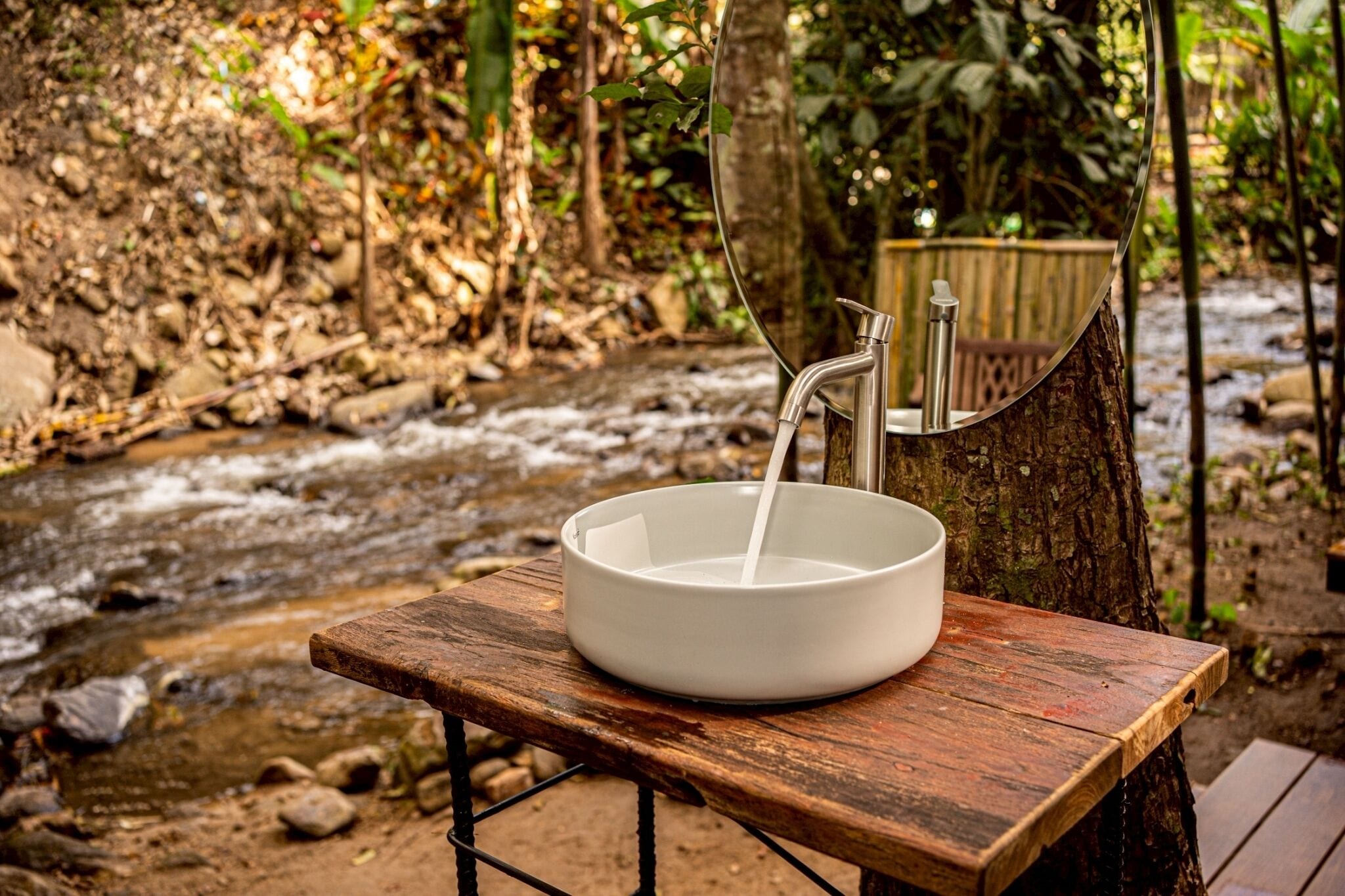 A white basin on a wooden table with a metal faucet, set against a backdrop of a stream and trees.
