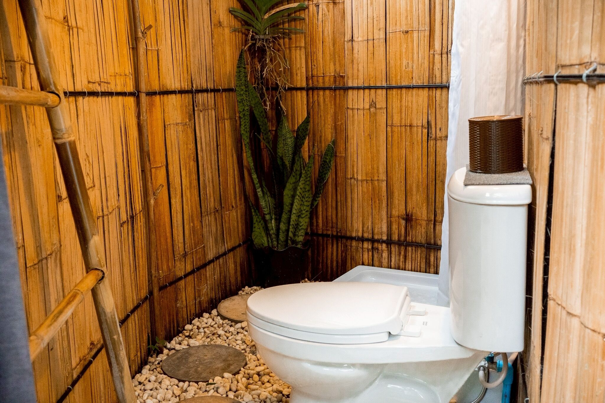 Une salle de bain aux murs recouverts de bambou, équipée de toilettes, d’un chauffe-eau et d’une plante.