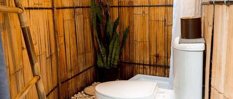 Une salle de bain aux murs recouverts de bambou, équipée de toilettes, d’un chauffe-eau et d’une plante.