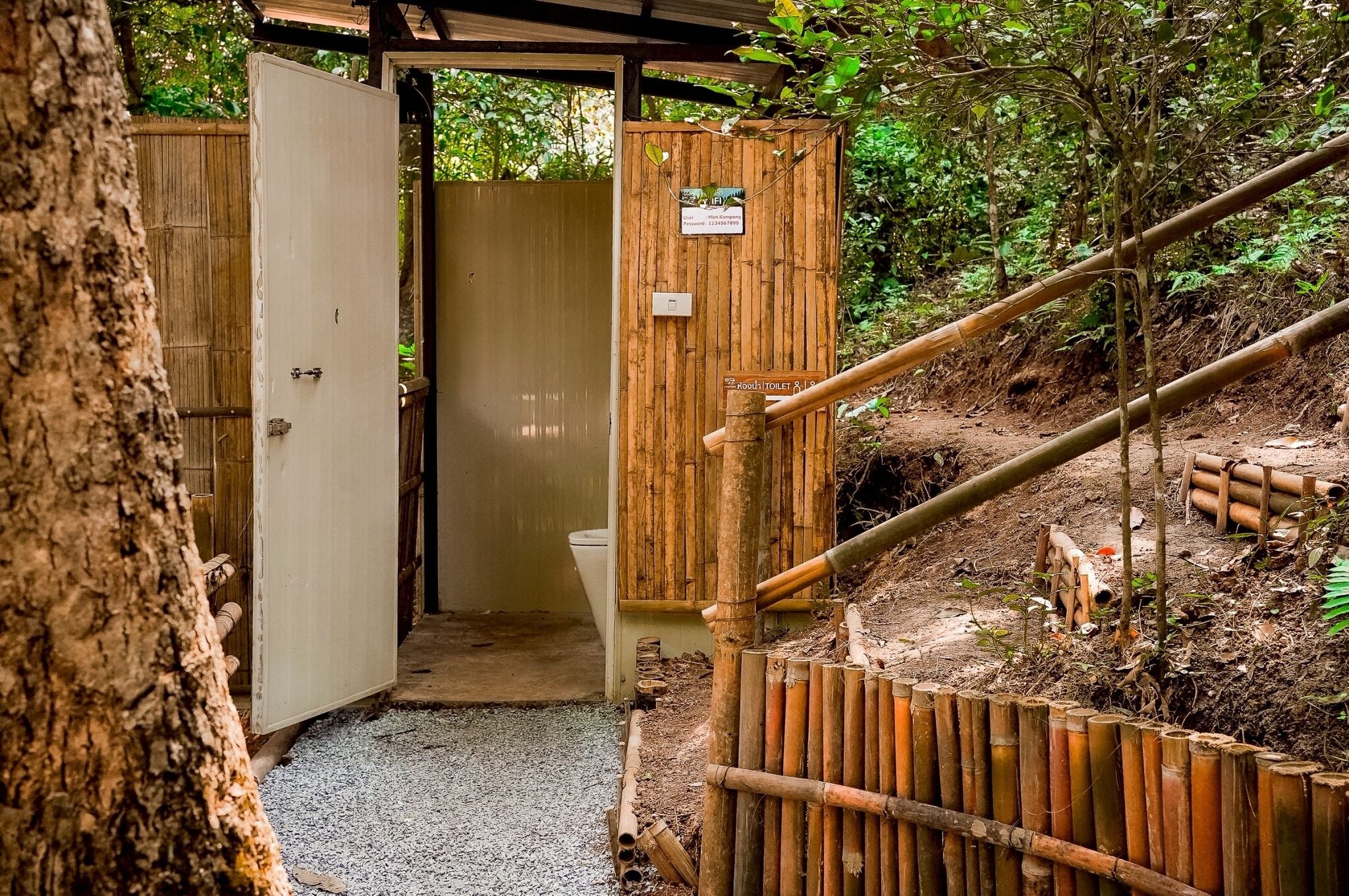 Un'ingresso in legno rustico con una porta che dà su una zona coperta, un segnale che indica i servizi igienici e uno sfondo naturale, con un bosco.