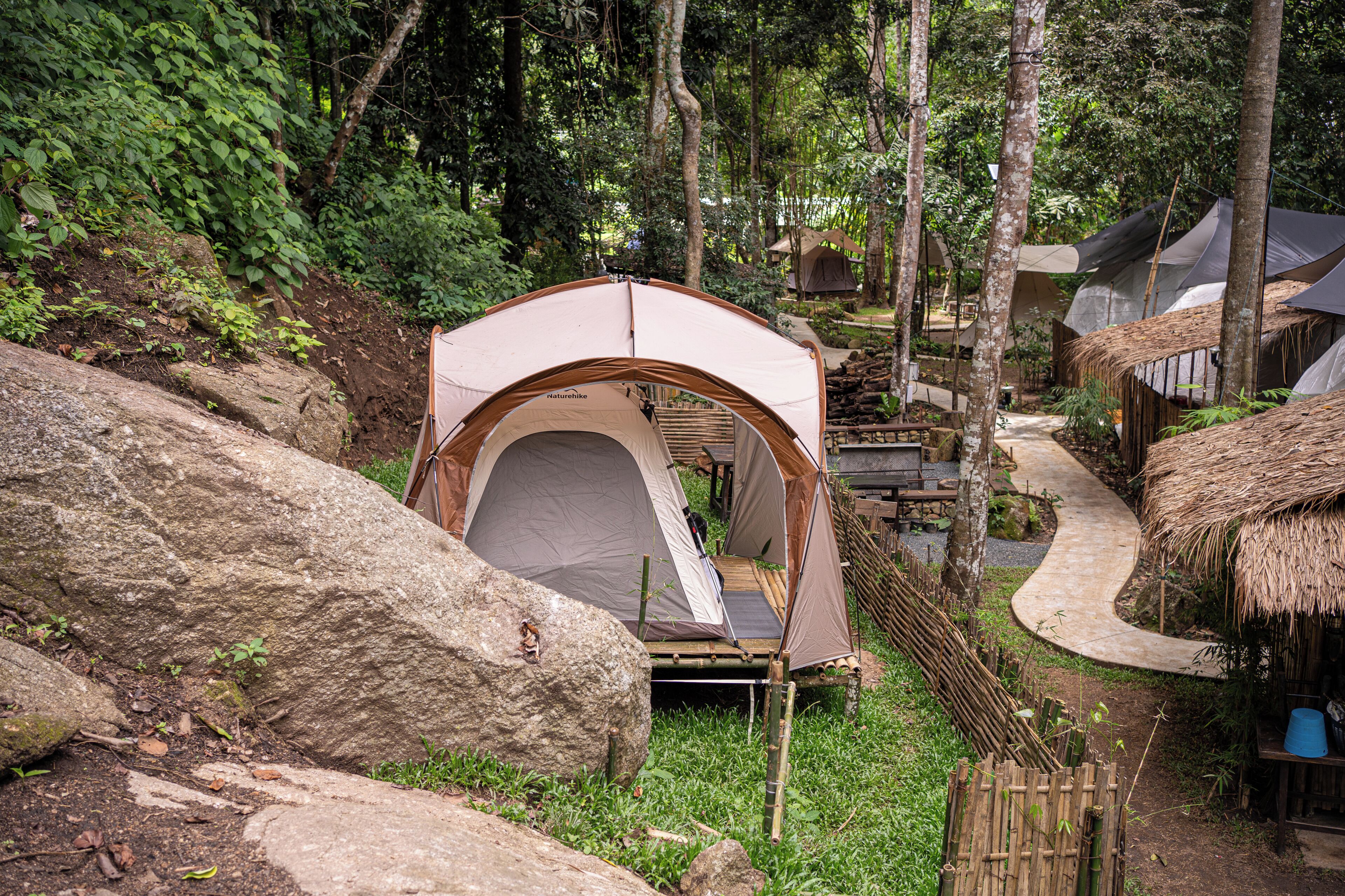 A nature-themed campsite with a single tent, surrounded by large rocks, bamboo fences, and a thatched-roof structure.