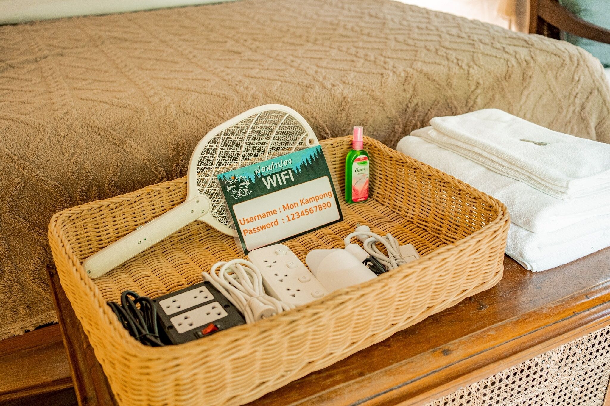 Ein Korbwägelchen mit einem WLAN-Symbol, einer Mehrfachsteckdose, einem Handyladegerät und einer kleinen Lotionflasche auf einem Bett.