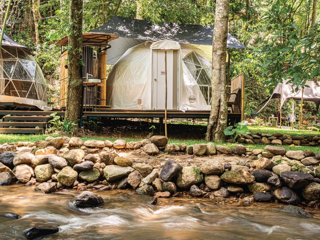 A rustic cabin with a wooden deck and a tent-like structure, surrounded by a natural landscape with a stream and rocks.