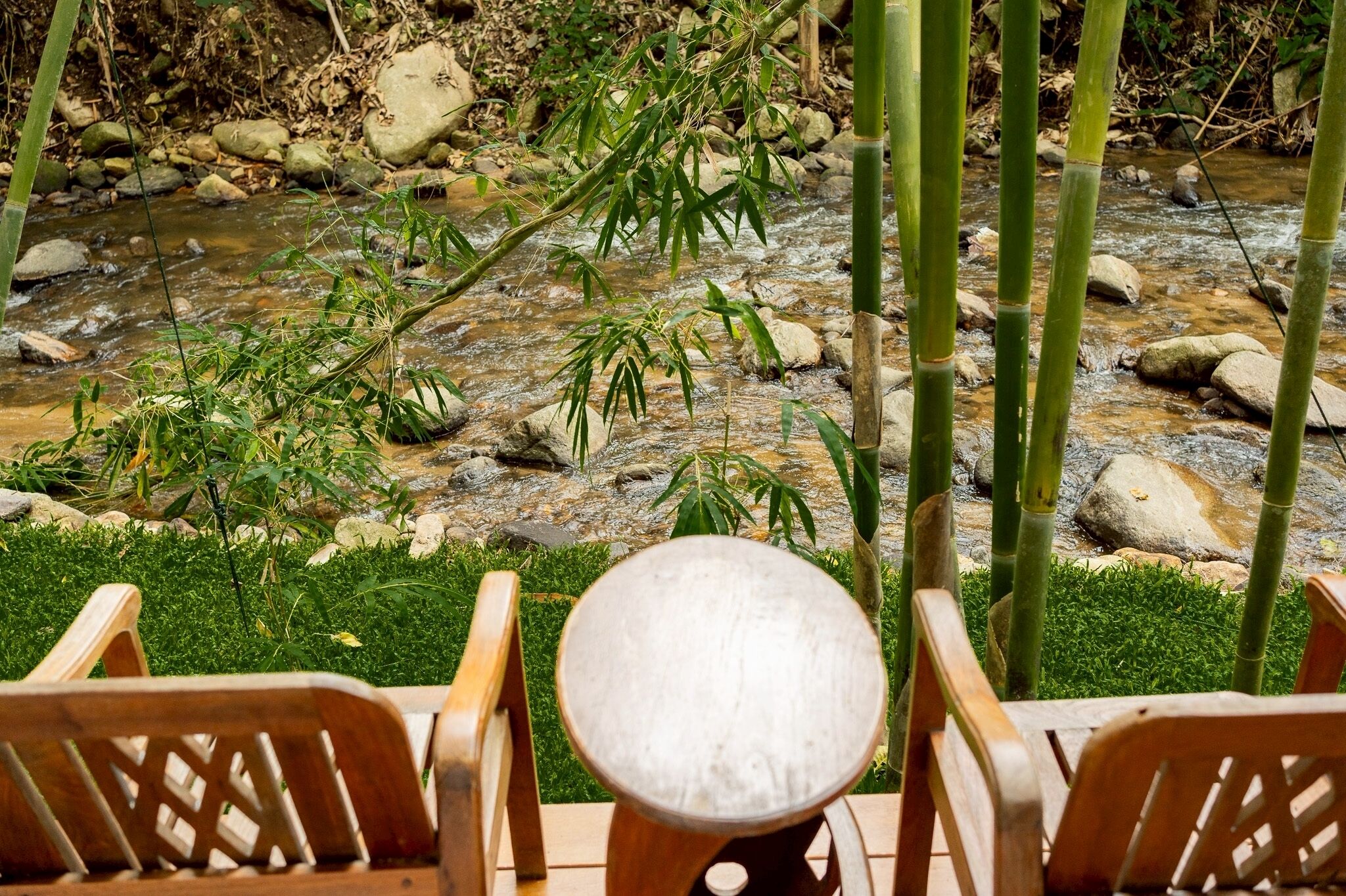 Mesa y sillas de madera junto a un arroyo con rocas y bambú.
