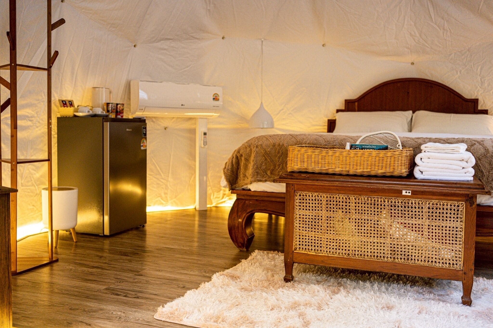 Ein Zeltraum mit einem Bett, einem Holztisch, einem Korbwägelchen und einem Kühlschrank.