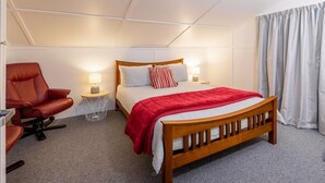 Ein Schlafzimmer mit einem hölzernen Bett, einem roten Sessel, einem Nachttisch mit Lampe und einem Vorhang.