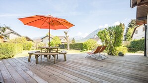 Een houten terras met een picknicktafel en een rode parasol, omringd door groen en huizen.