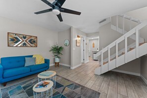 A modern living room with a blue sofa, a ceiling fan, a staircase, and a hallway leading to a kitchen. - Minutes to Old Town (Scottsdale)