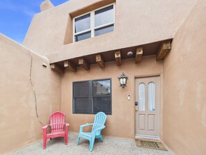 Een gebouw met een beige gevel, een houten veranda, een deur met een raam en twee stoelen.