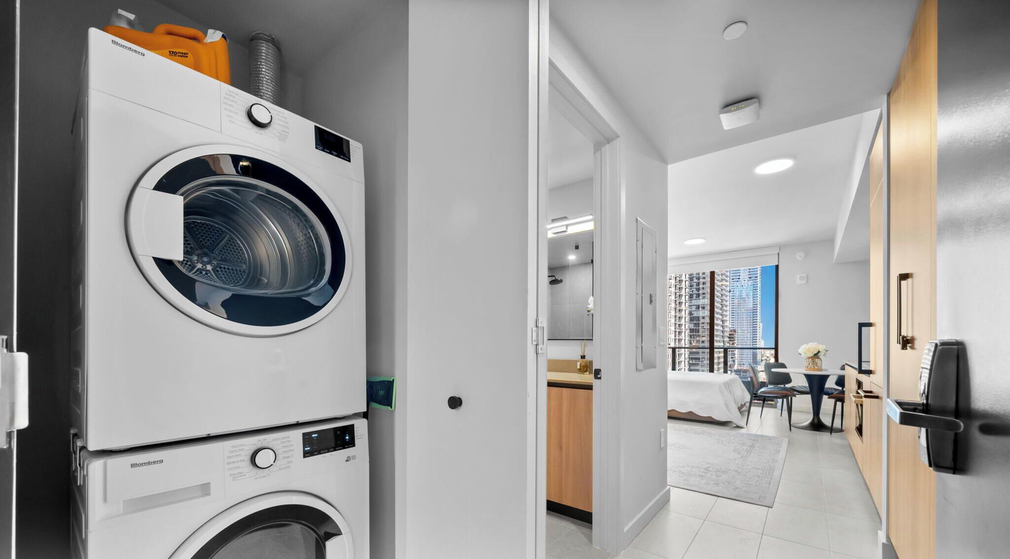 A modern apartment with a front-loading washing machine, a kitchenette, a dining area with a view, and a balcony.