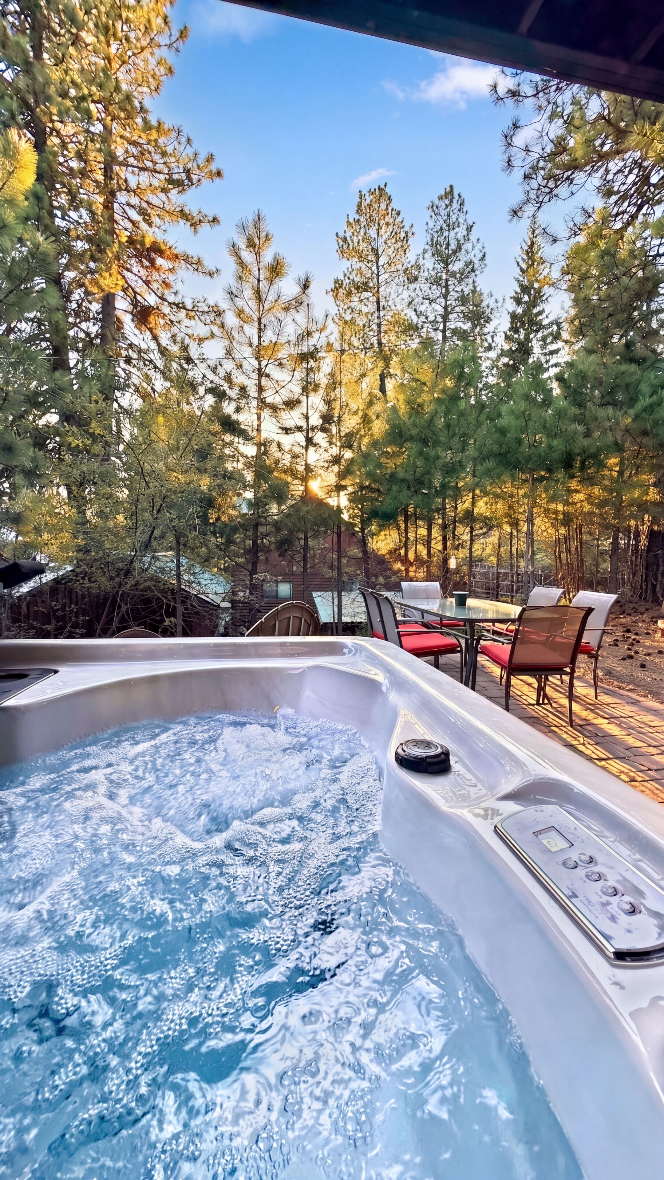 A hot tub with a control panel, surrounded by outdoor chairs and tables, set against a backdrop of trees and a clear sky.