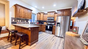 A modern kitchen with wooden cabinets, a central island with stools, stainless steel appliances, and a tiled backsplash. - The Timberline Haus - Where Pines Meet The Payette (McCall)