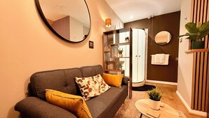 A modern living room with a sofa, a round mirror, a small table with a plant, and a shower area.