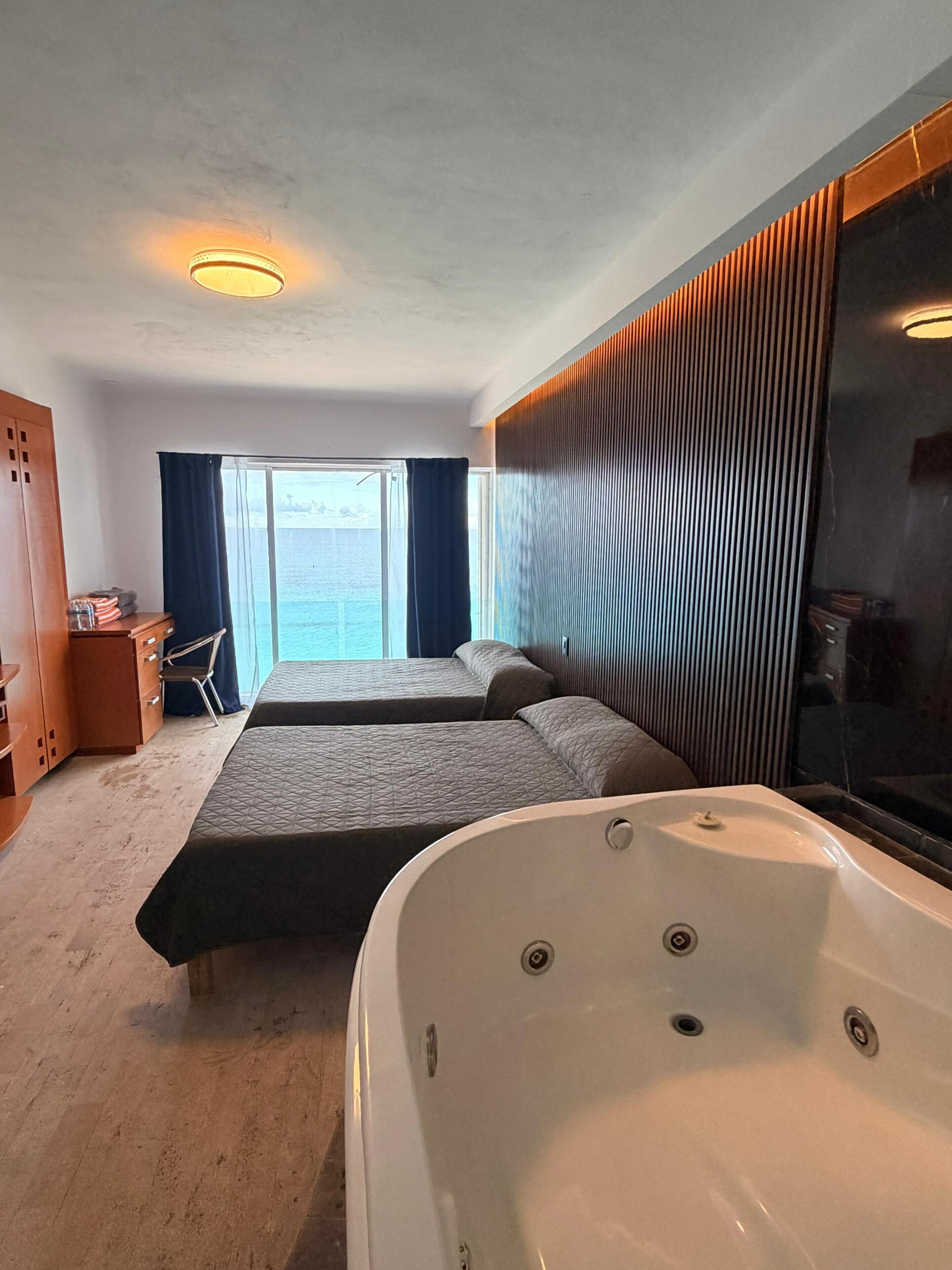 Ein Hotelzimmer mit zwei Betten, einer großen Badewanne, einem Holzschrank, einem Schreibtisch und Meerblick.