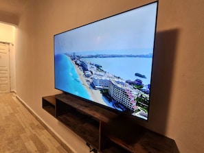 A flat-screen TV mounted on a wooden stand against a wall, displaying a coastal cityscape with buildings and a beach. - LUNA Y SOL CARIBE SUITES BY CANCUN PLAZA (Cancún)