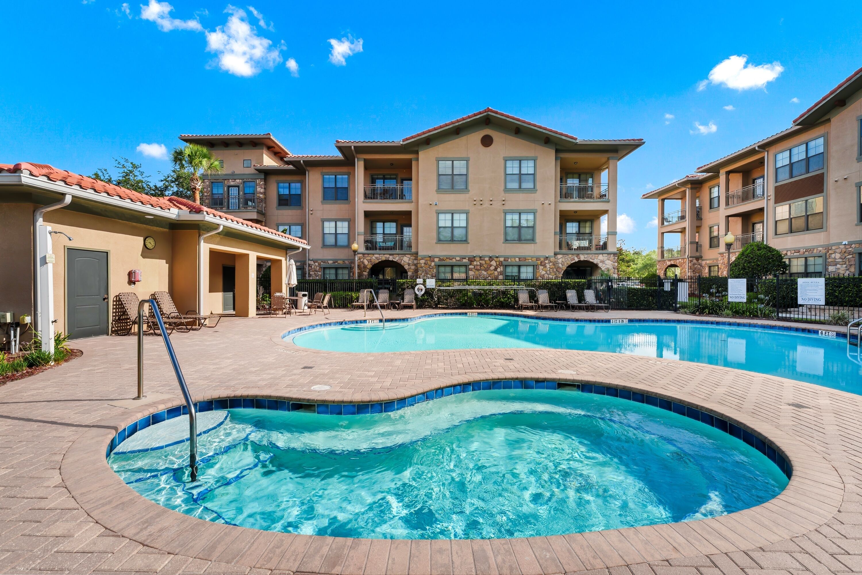 A swimming pool area with two pools, a covered patio, and a building with multiple balconies.