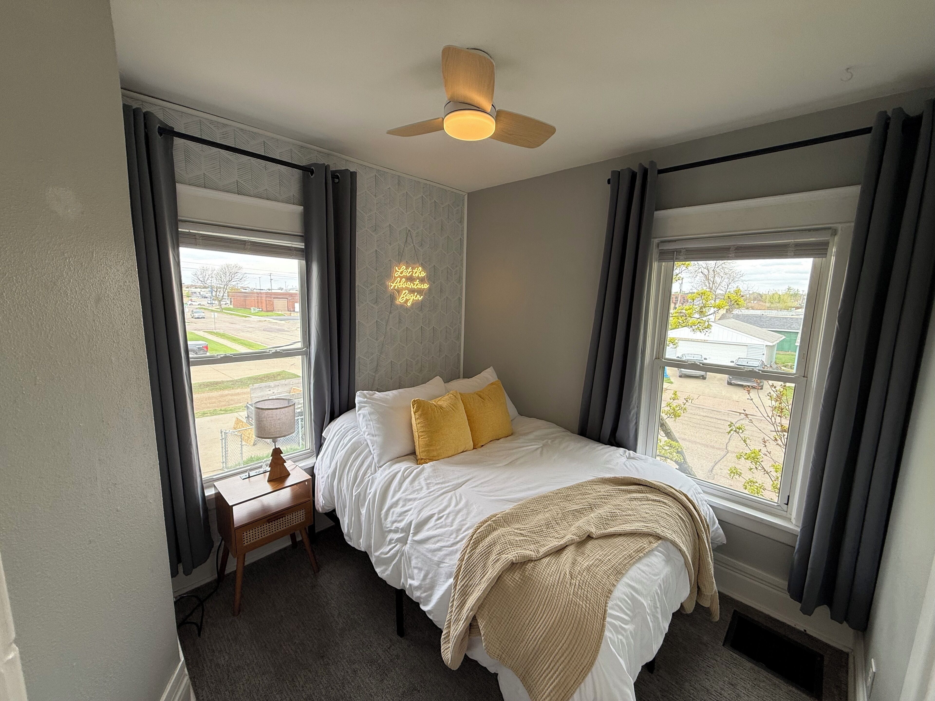 A bedroom with a bed, a desk, a chair, a ceiling fan, and large windows with curtains.