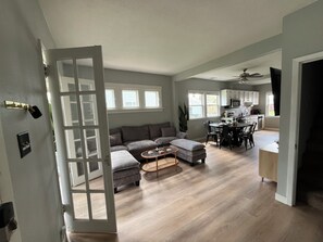 Amplia sala de estar con un sofá gris, un reposapiés y una mesa de centro, un comedor con sillas, y una cocina con armarios blancos y un ventilador de techo.