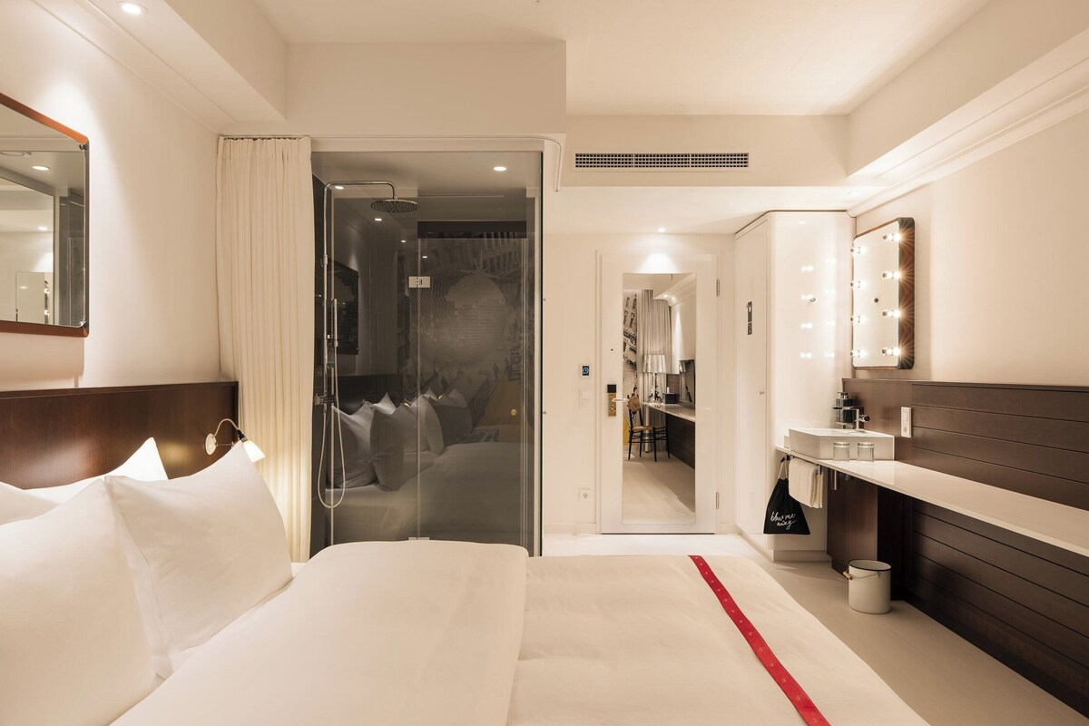 A hotel room with a bed, a bathroom with a mirror, and a hallway leading to a dining area.