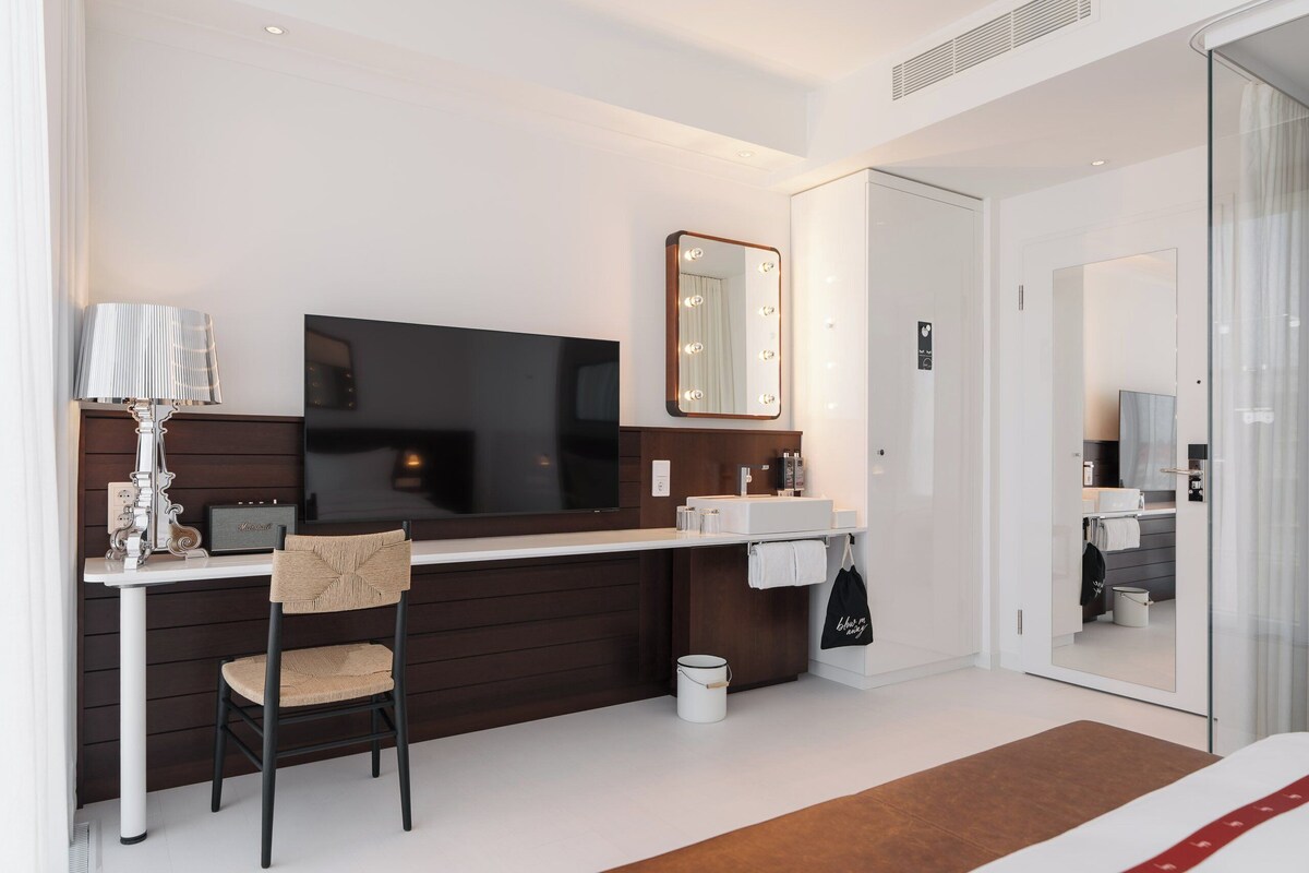 A modern hotel room with a flat-screen TV, a vanity with a mirror, a desk with a lamp, and a chair.