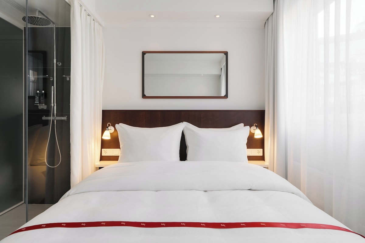 Habitación de hotel con cama, ducha, espejo y lámparas de noche.