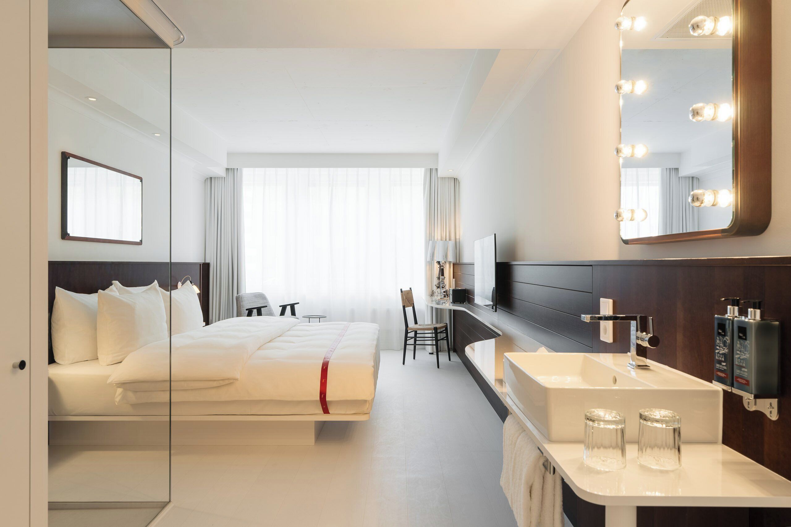 A hotel room with a bed, a desk, a mirror with lights, a sink, and a large window.