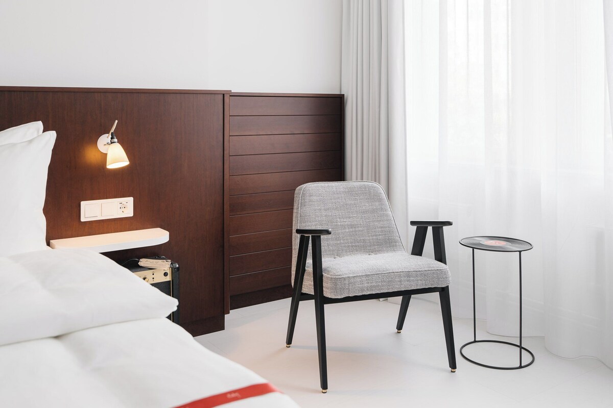 A hotel room with a bed, a chair, a small table, a wall-mounted light, and a window with curtains.
